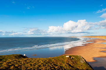 A scenic view from a grassy cliff overlooking a vast beach and ocean. The tide is out, revealing a wide expanse of sand, and the sky is a mix of blue and fluffy white clouds.