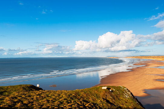 A scenic view from a grassy cliff overlooking a vast beach and ocean. The tide is out, revealing a wide expanse of sand, and the sky is a mix of blue and fluffy white clouds.