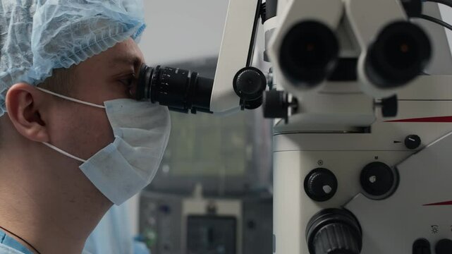 Side-view close-up of male ophthalmologist observing through lenses of surgical microscope during delicate eye procedure in sterile clinical environment