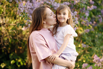 Fototapeta premium Loving mother kisses her smiling daughter on the cheek while holding her in a blooming lilac garden. Tender and joyful family moment in nature.
