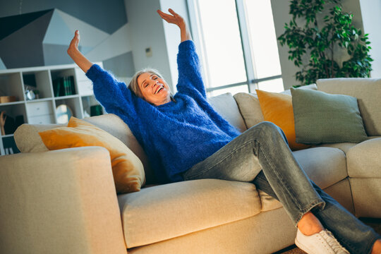 Smiling elderly woman in cozy sweater relaxing with arms raised on a comfortable couch at home during a sunny day - Powered by Adobe