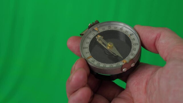 A close-up shot of a hand holding a compass, a vintage military-style compass with a metal casing and a glass cover over the dial. The compass features a sighting mechanism and a thumb loop