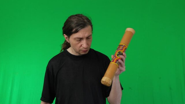 A man with a thoughtful expression holds and inspects a didgeridoo against a vibrant green screen, providing ample space for custom backgrounds