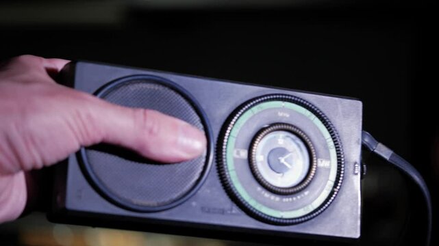 A hand holds a classic portable radio with a prominent tuning dial and speaker, set against a dark, moody background, suggesting nostalgia or a signal search