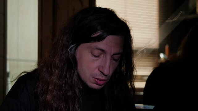 A man with long hair and a thoughtful expression, with his finger touching his lip, appears to be deeply contemplating or reacting to something unseen, possibly news
