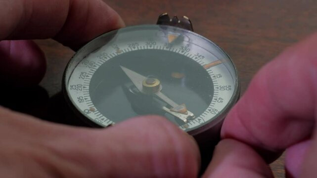 A close-up shot of a hand holding a compass, a vintage military-style compass with a metal casing and a glass cover over the dial. The compass features a sighting mechanism and a thumb loop