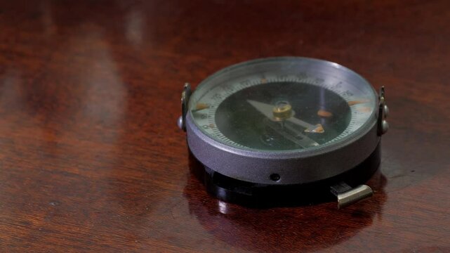 A close-up shot of a hand holding a compass, a vintage military-style compass with a metal casing and a glass cover over the dial. The compass features a sighting mechanism and a thumb loop