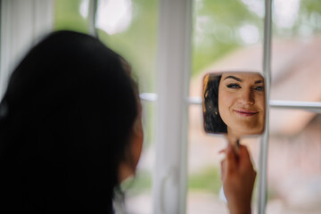 A smiling woman with elegant makeup admires her reflection in a handheld mirror by the window, surrounded by soft daylight and natural tones.. © Raivo