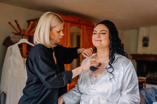 A makeup artist applies foundation on a bride’s chest as she prepares for her wedding day, wearing a white satin robe and smiling confidently..