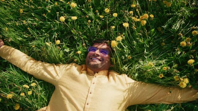Indian man wearing kurta pajama and sunglasses lying in dandelion field, experiencing moment of pure joy with open arms and radiant smile against vibrant natural backdrop