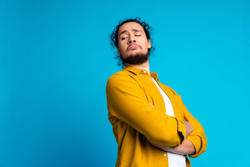 Confident young man in a stylish yellow shirt posing against a blue background with a playful...