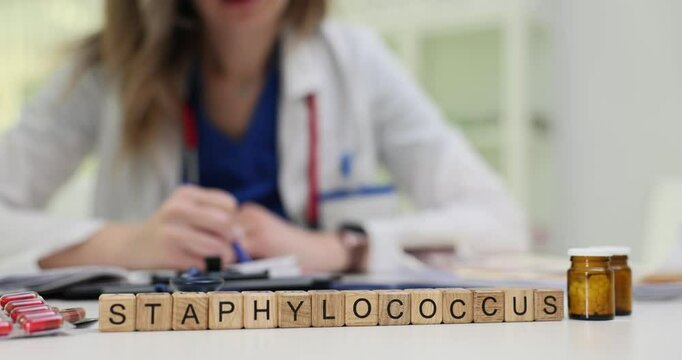 Female doctor writes prescription near wooden cubes with Staphylococcus and medication bottles on medical desk. Infection treatment on clinic setting
