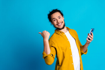 Cheerful young man in trendy yellow shirt holding smartphone and gesturing against blue background,...