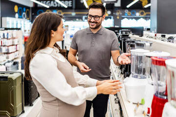 Smiling shop assistant helping a pregnant customer choose home appliances while showcasing various products in a vibrant electronics store