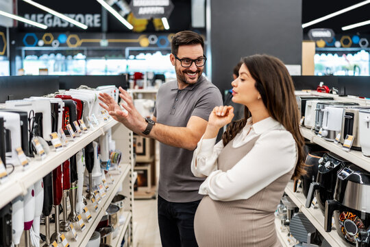 Smiling shop assistant helping a pregnant customer choose home appliances while showcasing various products in a vibrant electronics store