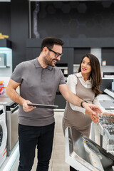 Smiling salesman holding a tablet, discussing and showing dishwashers to a pregnant customer, selecting the best options in a modern home appliance store