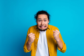 Excited young man in a yellow shirt expressing enthusiasm with clenched fists over a vibrant blue...
