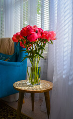 Bright pink peonies in a clear glass vase on a rustic wooden table, cozy interior with teal armchair, houseplants, and soft natural light. Homey and vibrant atmosphere.