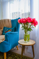 Bright pink peonies in a clear glass vase on a rustic wooden table, cozy interior with teal armchair, houseplants, and soft natural light. Homey and vibrant atmosphere.