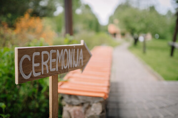 A wooden signpost with &ldquo;CEREMONIJA&rdquo; points to a garden path, suggesting the direction to a wedding or special event ceremony outdoors..