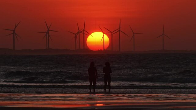 Two people admire sunset with wind turbines in Maceio, Brazil