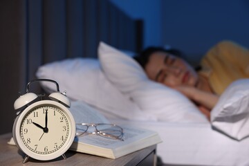 Young woman sleeping in bed at home, focus on alarm clock, glasses and book