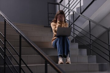 Cyber bullying. Upset teenage girl with laptop on stairs indoors