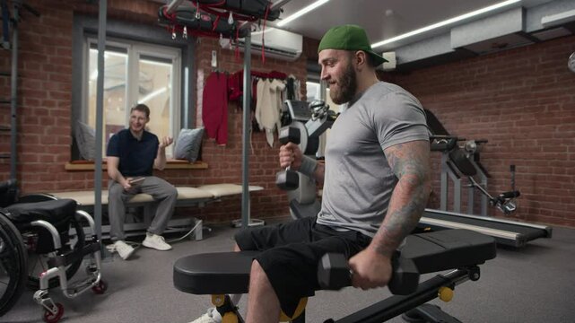 Tattooed bodybuilder with disability sitting on bench and doing bicep curls while his trainer holding tablet and telling funny story in blurred background