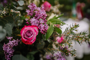 Close-up of a fresh pink rose with raindrops, surrounded by lilacs, greenery, and white blossoms in a vibrant floral arrangement..