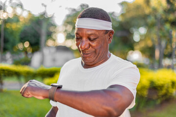 Senior black man in outdoor gym clothes and looking at smart watch. White clothes and blurred background.
