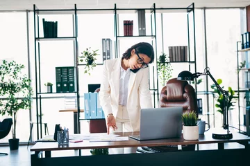 Gordijnen Confident female executive working in a modern office while multitasking with phone and laptop for professional productivity © deagreez