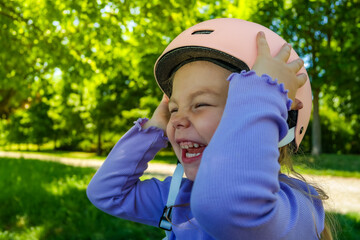 A little cute girl puts a pink helmet on her head and smile. She learns to roller skate or ride a...