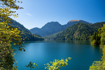 picturesque panorama: sunbeams break through the leaves of the trees, revealing a view of the beautiful Lake Ritsa, the heart of the Abkhazian mountain range. It is a warm autumn day.