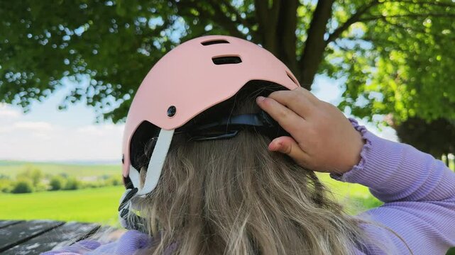 A little girl puts on and tightens a pink protective helmet on her head with her hand. Helmet size, summer sports.