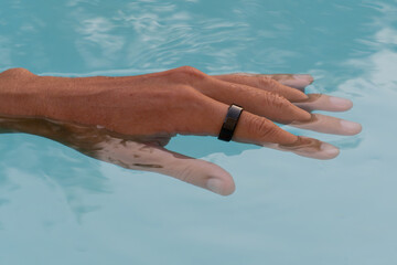 Hand, submerged in clear pool water showcasing a sleek waterproof black smart ring with health tracker.