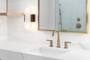 A bathroom detail with a subway tile backsplash, gold faucet and mirror, white marble countertop, and a modern sconce mounted on the wall.