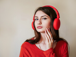 Attractive girl in red shirt with red big wireless headphones, light color wall background. Young...