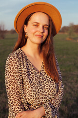 A smiling woman in a leopard print dress and hat poses outdoors in soft evening light.