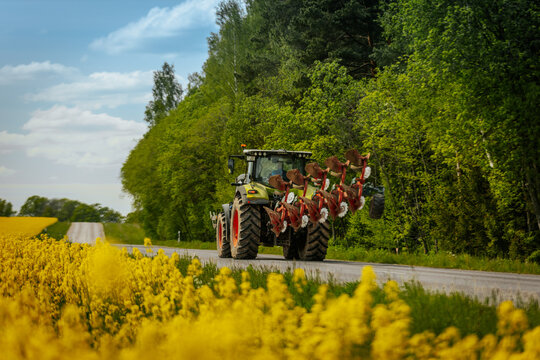 A large green tractor with red plow equipment drives along a rural road lined with blooming yellow flowers and lush green trees under a blue sky.
