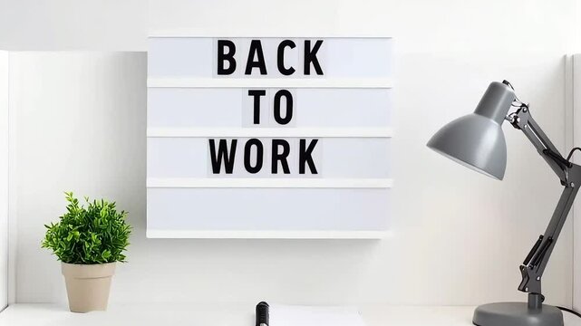 Back to work message on a lightbox on a minimalist white desk with a lamp and plant. Perfect for blog, social media, or website.