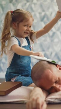 closeup of young bald bearded father sleeps on floor in room, small cheerful daughter wakes him up, hits him on head with notebooks, dad winces and falls asleep again girl with pigtails smiles laughs