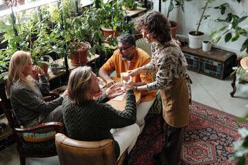 Group sitting around wooden table in cozy cafe surrounded by greenery, engaging in lively conversation and sharing tea while server hands small teacup to person wearing green sweater