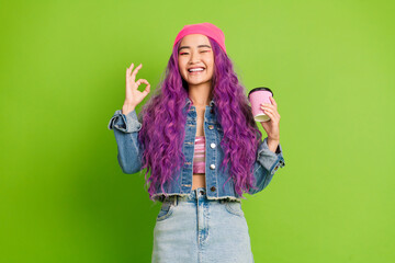 Young woman with purple hair wearing denim and making a hand gesture while sipping coffee against a...