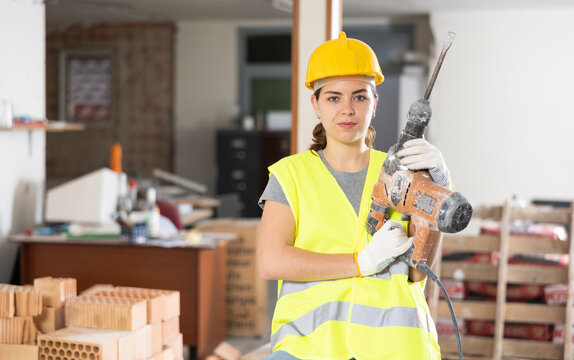 Portrait of young female builder wearing protective gear posing with handheld demolition hammer at indoor construction site