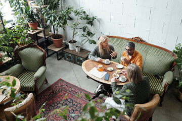 Overhead view of people sitting at round table, drinking coffee and conversing in cozy cafe with green plants and vintage furniture