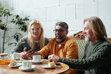 Senior friends sharing digital memories while having tea at cozy cafe. Elderly caucasian individuals seated around table with pastries, smartphone, and lively conversation taking place