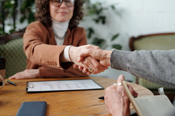 Professional setting with two people shaking hands across table, indicating agreement or partnership. Background includes business documents and planner on table