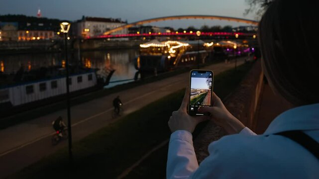 Over shoulder shot: tourist capturing scenic cityscape of illuminated bridge during sunset, using smartphone while standing on riverbank in slow motion. Tripper captures picturesque cityscape