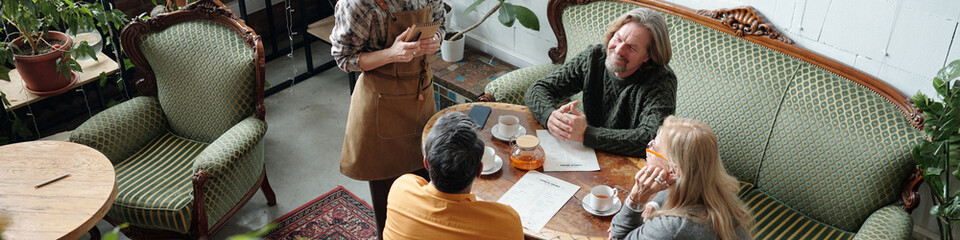 Group of people engaging in conversation while seated at a wooden table in cozy cafe setting. Green upholstered chairs and plants fill the space, creating inviting atmosphere