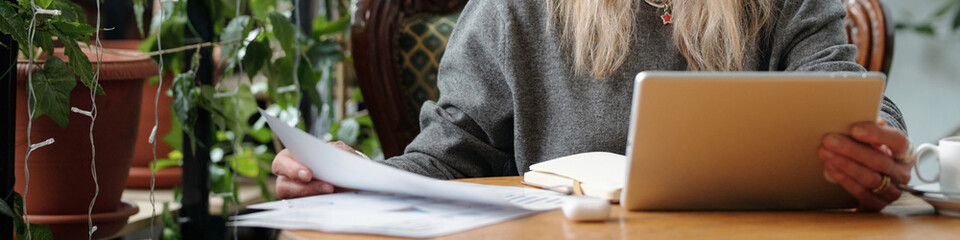 Woman sitting at table reviewing papers and using tablet in cozy environment surrounded by plants. Casual attire and focused expression while working